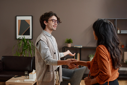 Horizontal medium shot of young psychologist greeting woman shaking hands and offering her to take seat