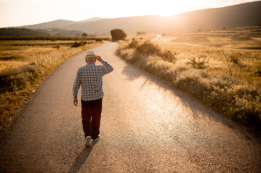 Back view of senior man with hat while walking on country road on sunny day