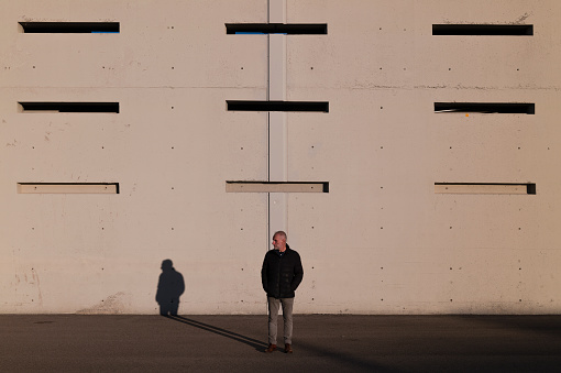Full length of adult man in winter clothes standing against white wall of a stadium in Madrid, Spain, with sunlight and shadow.