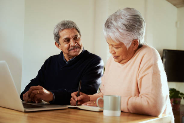 istock Multi-cultural elderly couple sitting at modern kitchen counter with laptop, wife making notes in notebook. Budgeting for retirement 1359480255