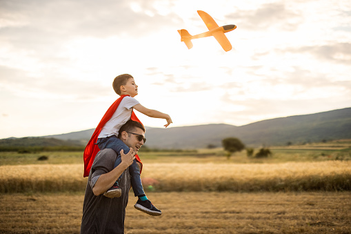 Happy father carrying on shoulders his son while they playing with airplane toy in nature