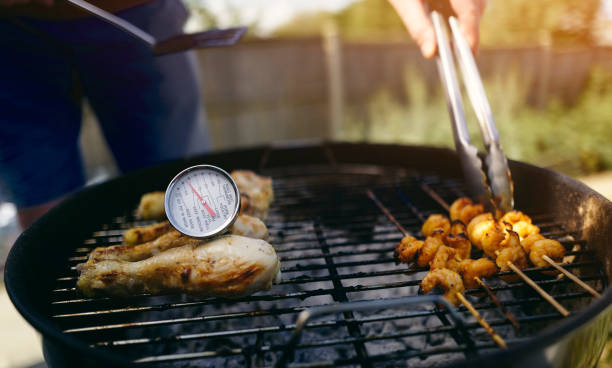 A food thermometer checking that chicken is being cooked correctly, safely on a barbecue as a man turns over prawns with a pair of tongs on a sunny day in the garden.