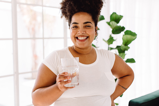 Cheerful plus size woman with glass of water smiling at camera. Healthy female holding glass of water.