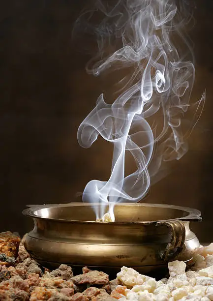 Frankincense and Myhrr burn with curling smoke patterns in an antique brass bowl. Chunks of Frankincense and Myhrr are in the foreground.