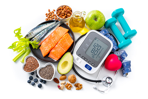 Overhead view of a white background filled with multicolored healthy food. A blood pressure monitor, stethoscope and dumbbells complete the composition. The composition includes salmon, sardines, green apple, nuts, blueberries, olive oil, dates, celery, avocado, broccoli, pinto beans, chocolate, flax seeds among others. High resolution 42Mp studio digital capture taken with SONY A7rII and Zeiss Batis 40mm F2.0 CF lens