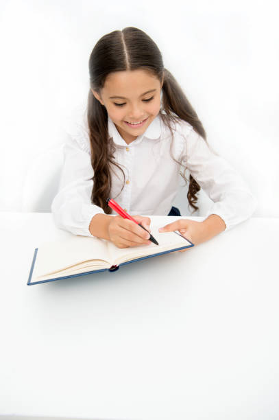 istock Little bookworm. Writing notices. Girl child writes hometask while sit table white background. Schoolgirl studying writing notice. Kid girl school uniform happy face writes exercise, copy space 1359487428