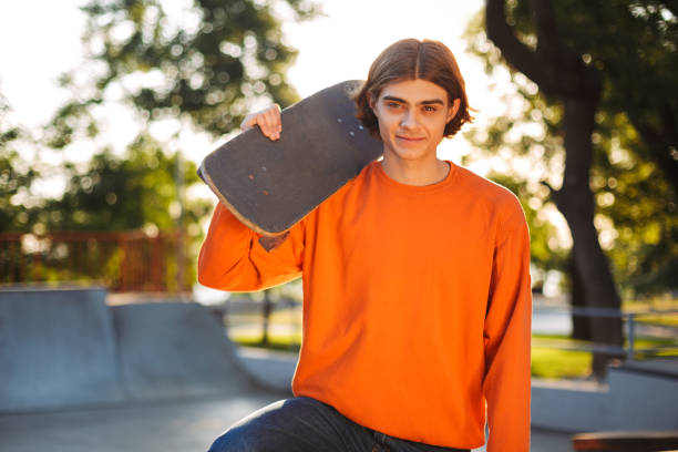 istock Young smiling skater in orange pullover happily looking in camera while holding  skateboard on shoulder with skatepark on background 1359502663