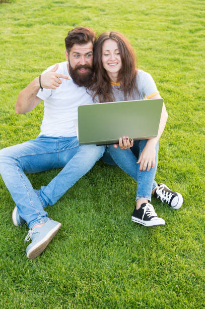 happy couple use laptop on green grass. work outdoors benefits. working on computer in park. man and woman surfing internet on notebook. students life. online shopping. communication online - surfing wireless vertical outdoors lifestyles imagens e fotografias de stock