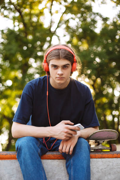 istock Young guy in orange headphones holding cellphone in hand thoughtfully looking in camera while spending time at modern skatepark 1359503880