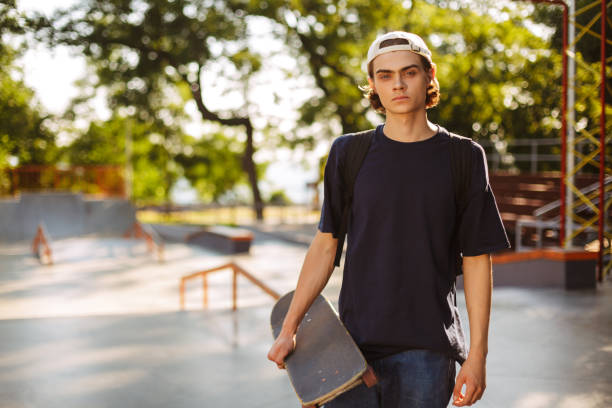 Young serious guy in black T-shirt and white cap thoughtfully looking in camera holding skateboard in hand with modern skatepark on background