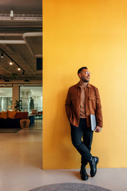 Creative businessman looking away in a modern office Creative young businessman looking away with a smile while standing in a co-working space. Happy young businessman holding a laptop and leaning against the wall in a modern office. looking away stock pictures, royalty-free photos & images