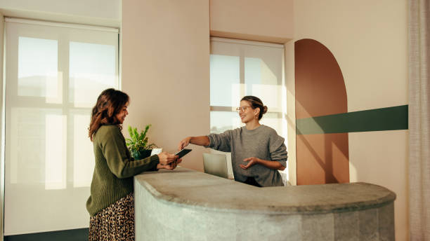 Carefree receptionist assisting a woman with signing in to an office Carefree receptionist assisting a woman with signing in to an office. Friendly receptionist showing a woman where to sign on a digital tablet. Woman working at the front desk of a co-working space. hotel reception stock pictures, royalty-free photos & images