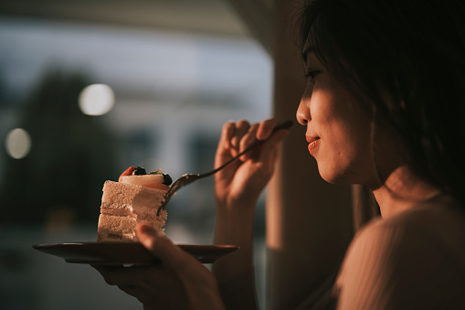 Asian Chinese beautiful woman enjoying slice of cake at home during sunset looking away through window curtain