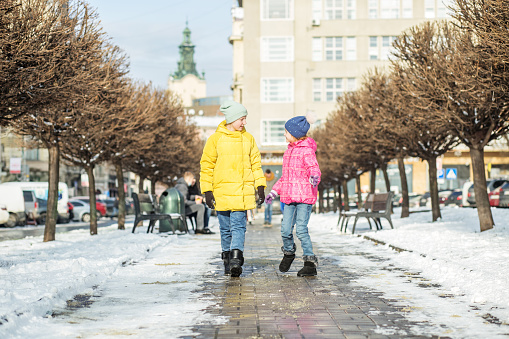 Two children talk and walk down the street in winter jackets and warm hats. Winter season. Beautiful city. A happy family. Cheerful little sisters.