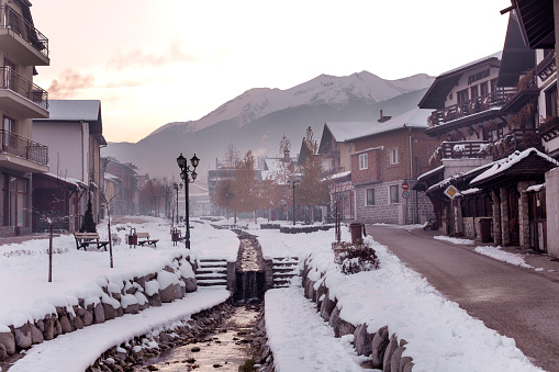 Bansko, Bulgaria - December 6, 2019: Gotse Delchev street view with old, traditional houses, water stream and Pirin mountains