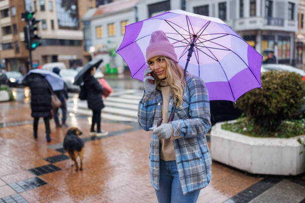 istock Young woman using phone while standing on sidewalk during rainy day at city 1359516277