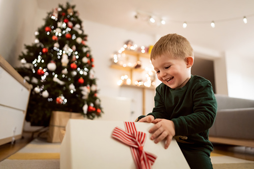 Caucasian toddler boy sitting near Christmas tree, and unwrapping his Christmas present