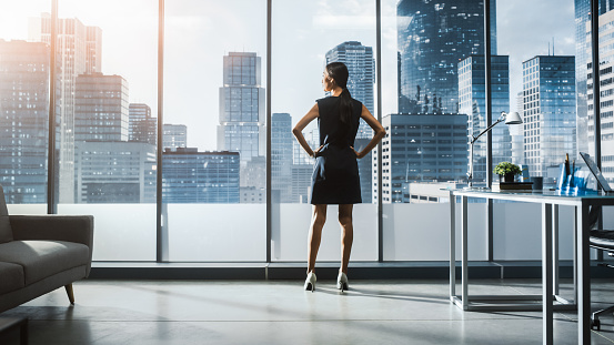Successful Businesswoman in Stylish Dress Looking out of the Window at Big City in Downtown Area. Confident Female CEO Working on Financial Projects. Manager at Work Planning Marketing Campaign.