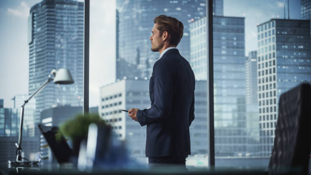 istock Successful Young Businessman in a Suit Standing in His Modern Office, Using Smartphone, Looking out of the Window on Big City with Skyscrapers. Successful Finance Manager Planning Work Projects. 1359523287