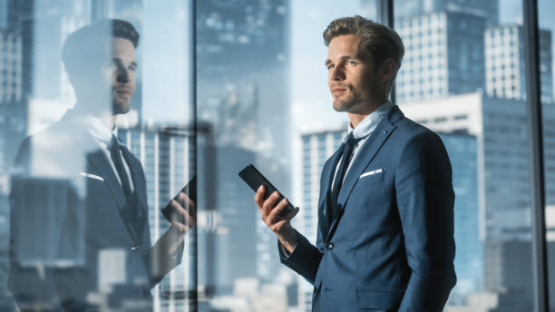 istock Confident Young Businessman in a Suit Standing in Modern Office, Using Smartphone, Looking out of the Window on Big City with Skyscrapers. Successful Finance Manager Planning Work Projects. 1359524122