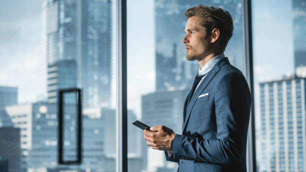 istock Confident Young Businessman in a Suit Standing in Modern Office, Using Smartphone, Looking out of the Window on Big City with Skyscrapers. Successful Finance Manager Planning Work Projects. 1359524123