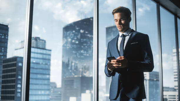 istock Confident Young Businessman in a Suit Walking in Modern Office, Using Smartphone, Looking out of the Window on Big City with Skyscrapers. Successful Finance Manager Planning Work Projects. 1359524136