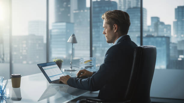 istock Confident Businessman in a Suit Sitting at a Desk in Modern Office, Using Laptop Computer, Next to Window with Big City with Skyscrapers View. Successful Finance Manager Planning Work Projects. 1359524354