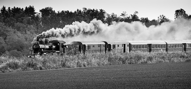 Vintage steam train with ancient locomotive and old carriages runs on the tracks in the countryside. Black and white photography.
