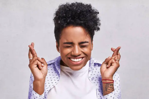 Photo of Beautiful African American female student crosses fingers with big hope, has positive expression, poses against the blank background. Dark skinned lovely young woman prays for good luck during exam