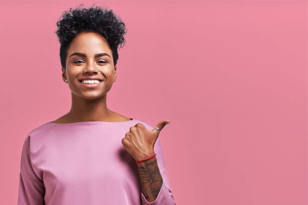 istock Omg concept. Joyful dark skinned female with surprised expression, smiled widely, points aside with thumb, shows something good on pink background. Advertisement concept 1359535833