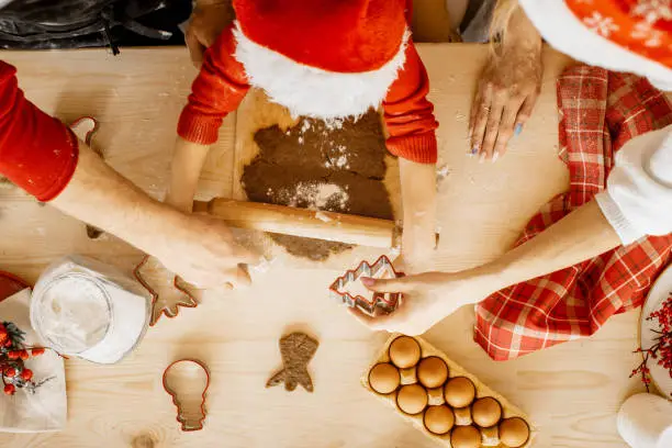Top view and close up of small hands roll dough holding a rolling pin. Mother is holding a tree shaped metal cutter and father helps to hold the cutting board. Christmas cookies and holidays.
