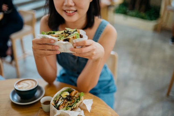 Young asian woman eating sandwich in cafe Image of a smiling Asian Chinese woman having toasted sandwich and latte in a cafe sandwich healthy lifestyle healthy eating bread stock pictures, royalty-free photos & images