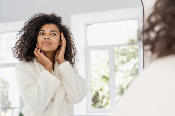 Young african american woman looking at mirror in bathroom Skincare, dermatology and cosmetology concept. Young african american woman standing at bathroom, looking at mirror. Female in bathrobe checking skin, touching her perfect face and smiling nice looking in mirror stock pictures, royalty-free photos & images