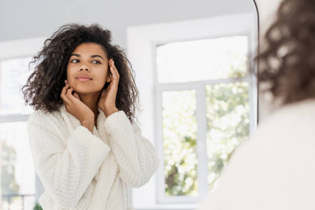 young african american woman looking at mirror in bathroom - mirror women bathroom make up imagens e fotografias de stock