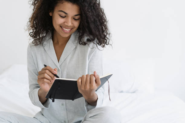 Dark skinned woman smiling wide, writing in diary Happy dark skinned woman smiling wide, writing notes in personal diary journal, while she sitting in bedroom wearing in pajamas. Inspiration, imagination and creativity concept personality test stock pictures, royalty-free photos & images