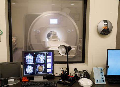 The control room of an fMRI machine.  You can see various images of a brain on the computer monitor in the foreground and the fMRI machine in the room beyond the glass.  No people.