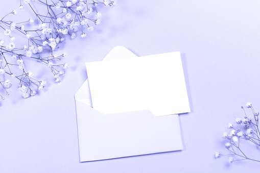 Pastel festive layout with envelope, empty sheet, white gypsophila flowers in color of year 2022 Very Peri. Holiday Mock up. View from above. Copy space for text.