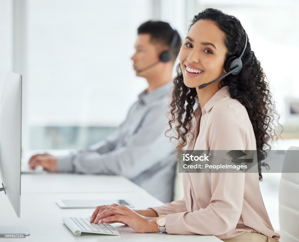 Portrait of a young call centre agent working on a computer in an office with her colleague in the background Just going about the day's calls Call Center Stock Photo