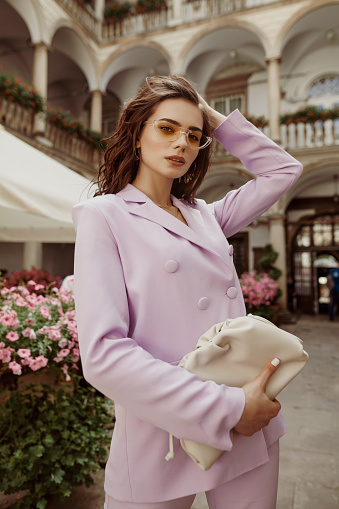 Outdoor fashion portrait of elegant woman wearing lilac suit, yellow sunglasses, holding trendy leather pouch handbag, posing in street of European city