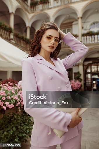 istock Outdoor fashion portrait of elegant woman wearing lilac suit, yellow sunglasses, holding trendy leather pouch handbag, posing in street of European city 1359562499