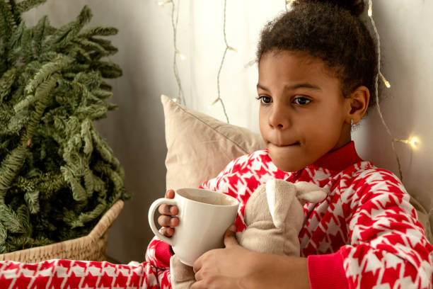 Thoughtful African-American girl in red and white pajamas is lying at home in bed and drinking cocoa.Fir branches in the corner of the bedroom and garlands on the wall.New Year and Christmas concept.