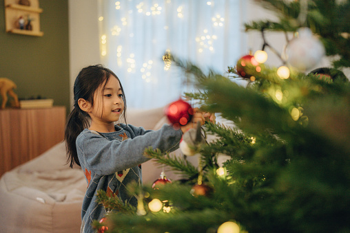 Joyful attractive family with little cute daughter decorating Christmas tree at home. Cheerful girl helping smiling parents to decorate xmas tree in living room during winter holidays.