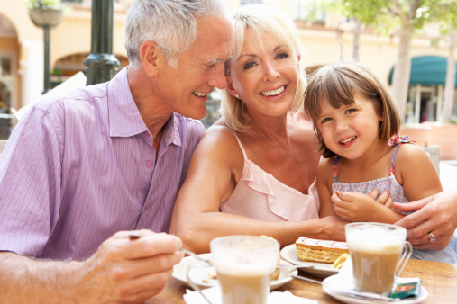 Grandparents With Granddaughter Enjoying Coffee And Cake Together In Caf