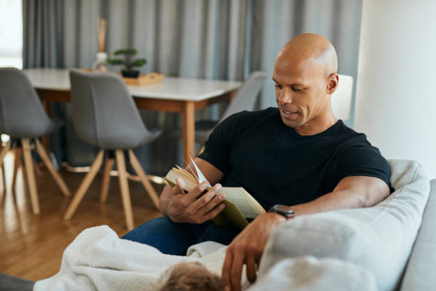 istock Young African American man reading book while relaxing on the sofa. 1359569284