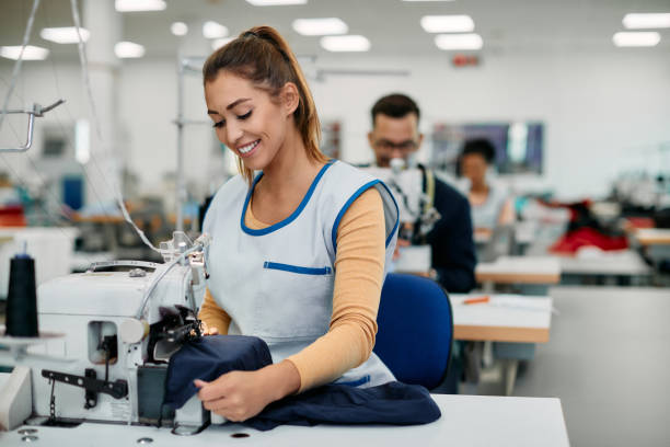 lavoratrice felice che cuce mentre lavora alla linea di produzione nella fabbrica di abbigliamento. - sewing tailor sewing machine women foto e immagini stock