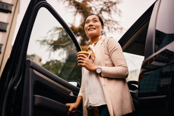 Below view of smiling Asian woman getting out of her car. Low angle view of Asian businesswoman with takeaway coffee getting out of a car. disembarking stock pictures, royalty-free photos & images