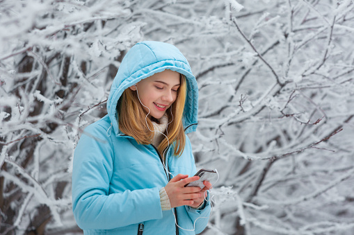Photo of a cute smiling young female watching videos online in a smartphone or listening to music with headphones while walking in a hood in a snowy winter park