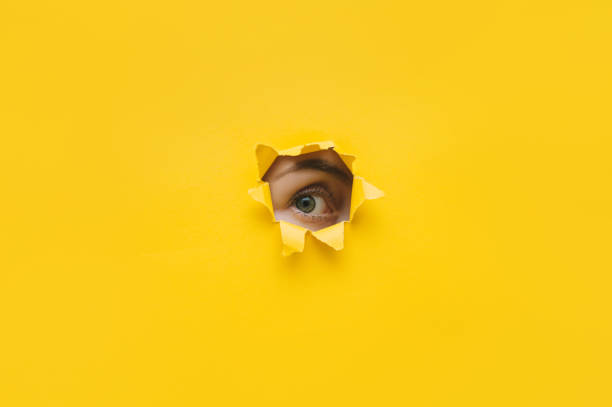 One eye looking through a hole in a yellow paper. Voyeurism. Woman is watching the husband. A curious look. Jealousy, spying on or overhearing the concept. Copy space. stock photo
