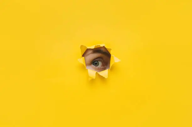 One eye looking through a hole in a yellow paper. Voyeurism. Woman is watching the husband. A curious look. Jealousy, spying on or overhearing the concept. Copy space.