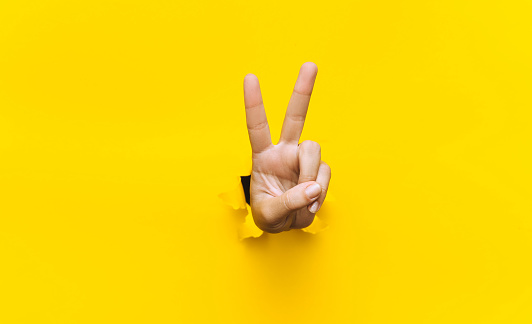 Victory sign two-finger hand gesture on yellow background with torn paper hole and copy space. It is ok concept.